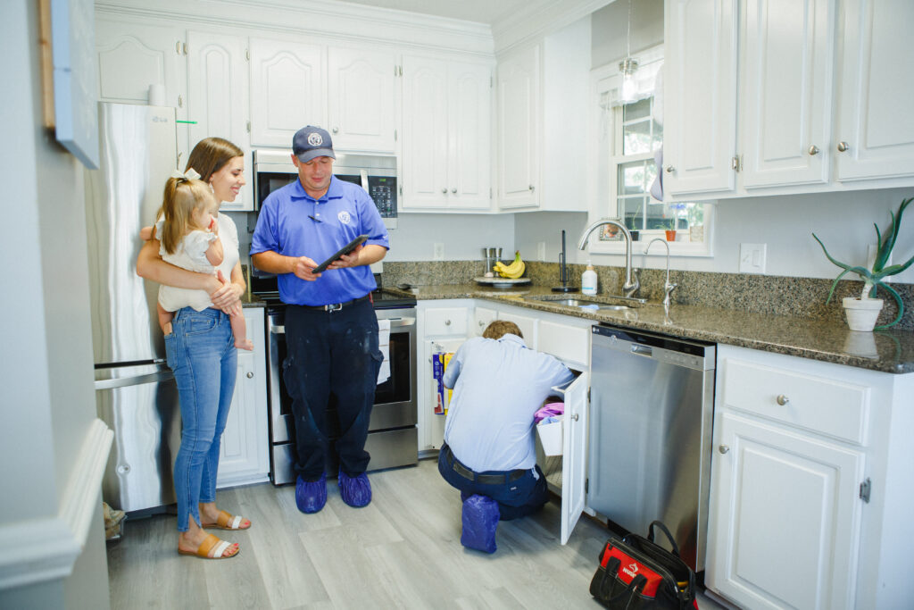 A Meetze plumbing technician holding an iPad in a kitchen while talking to a woman who is holding a female toddler. A second technician is working on the pipes underneath the kitchen sink. 